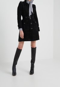 Vrouw in een met een riem omklede zwarte knopenjas met grote zakken, gestreepte shirt, gestreepte halsdoek en zwarte knielange leren laarzen.