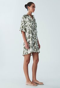 Sukienka na guziki wykonana z lekkiej tkaniny, z abstrakcyjnym zielonym wzorem na beżowym tle. Krótkie rękawy i boczne rozcięcia.