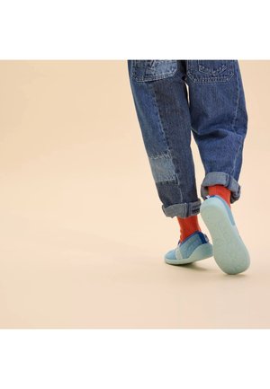 Enfant portant un jean à patchwork retroussé, des chaussettes orange vif et des chaussures légères bleu clair, marchant sur un sol beige.