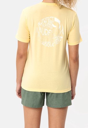 Person iført gul t-shirt med hvidt naturtema cirkelprint og grønne shorts, vist bagfra.