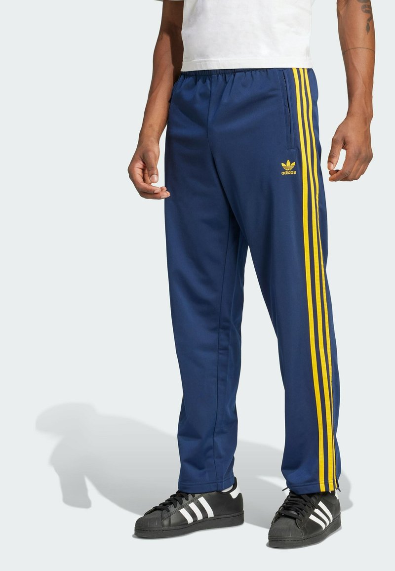 Blå träningsbyxor med gula ränder, elastisk midja, fickor och en Adidas-logotyp på vänster höft. Matchat med svarta sneakers.