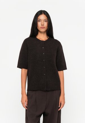 Woman with long dark hair wearing a buttoned, short-sleeve, dark brown sweater and loose dark brown pants, standing against a white background.
