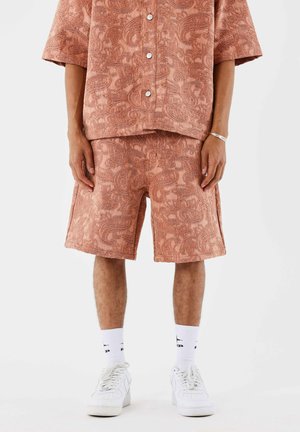 Homme portant une chemise et un short terracotta texturés assortis, avec des chaussettes blanches et des baskets, debout devant un fond blanc.