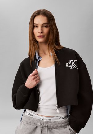 Junge Frau mit langen braunen Haaren, die eine schwarze, kurze Reißverschlussjacke mit „cK“-Logo, ein weißes Tanktop, graue Jogginghosen und einen sichtbaren Calvin Klein Bund trägt.