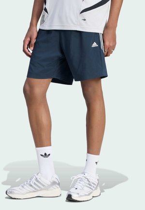 Personne portant un short Adidas bleu marine, des chaussettes Adidas blanches et des baskets Adidas blanches, debout devant un fond clair uni.