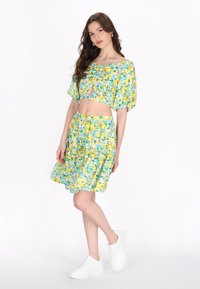 Virágmintás crop top és szoknyaszett, amely sárga, zöld és kék mintákkal díszített. Könnyű anyagból készült, lebegő kialakítással és feszes derékkal. Fehér sportcipő.
