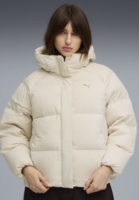 Giacca imbottita beige con colletto alto e cappuccio. Presenta una chiusura con zip, maniche larghe e un piccolo logo sul petto. Texture morbida.