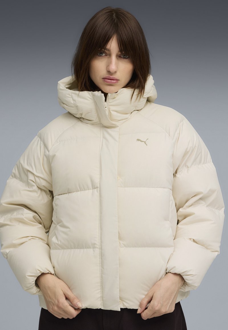 Beige vadderad jacka med hög krage och huva. Har dragkedja fram, vida ärmar och en liten logotyp på bröstet. Mjuk textur.