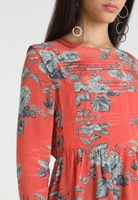 Koralowa sukienka w kwiatowy wzór, z długimi rękawami, marszczoną talią, teksturowanym korpusem, z szarymi i zielonymi akcentami kwiatowymi.