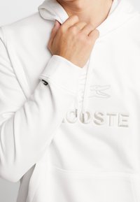 Vit hoodie med texturerat tyg, har en präglad logotyp och huva med dragsko. Muddar och fåll har en ren finish.