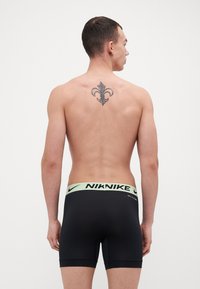 Črne bokserice z lahkono zelenim Nike pasom. Imajo gladko teksturo in prilegajoč dizajn. Tetovaža je vidna na hrbtu.