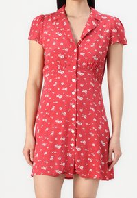 Rotes Blumen Kleid mit kurzen Ärmeln, Knopfverschluss vorne und weichem Stoff. Es hat ein tailliertes Oberteil und einen fließenden Rock. Kleine weiße Blumenmuster.