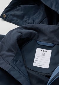 Tummansinisellä takilla on sileä pinta, vetoketjukiinnitys ja sisällä koko 116 ilmoittava etiketti. Mukana on nimikointi-osio henkilökohtaista käyttöä varten.