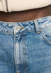 Jean en denim bleu clair avec une finition délavée, présentant des coutures orange, une fermeture à bouton et une étiquette de marque étiquetée "MULTPLY" sur la ceinture.