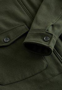 Dark green wool coat close-up showing sleeve cuff with black button and stitched pocket flap on textured fabric.
