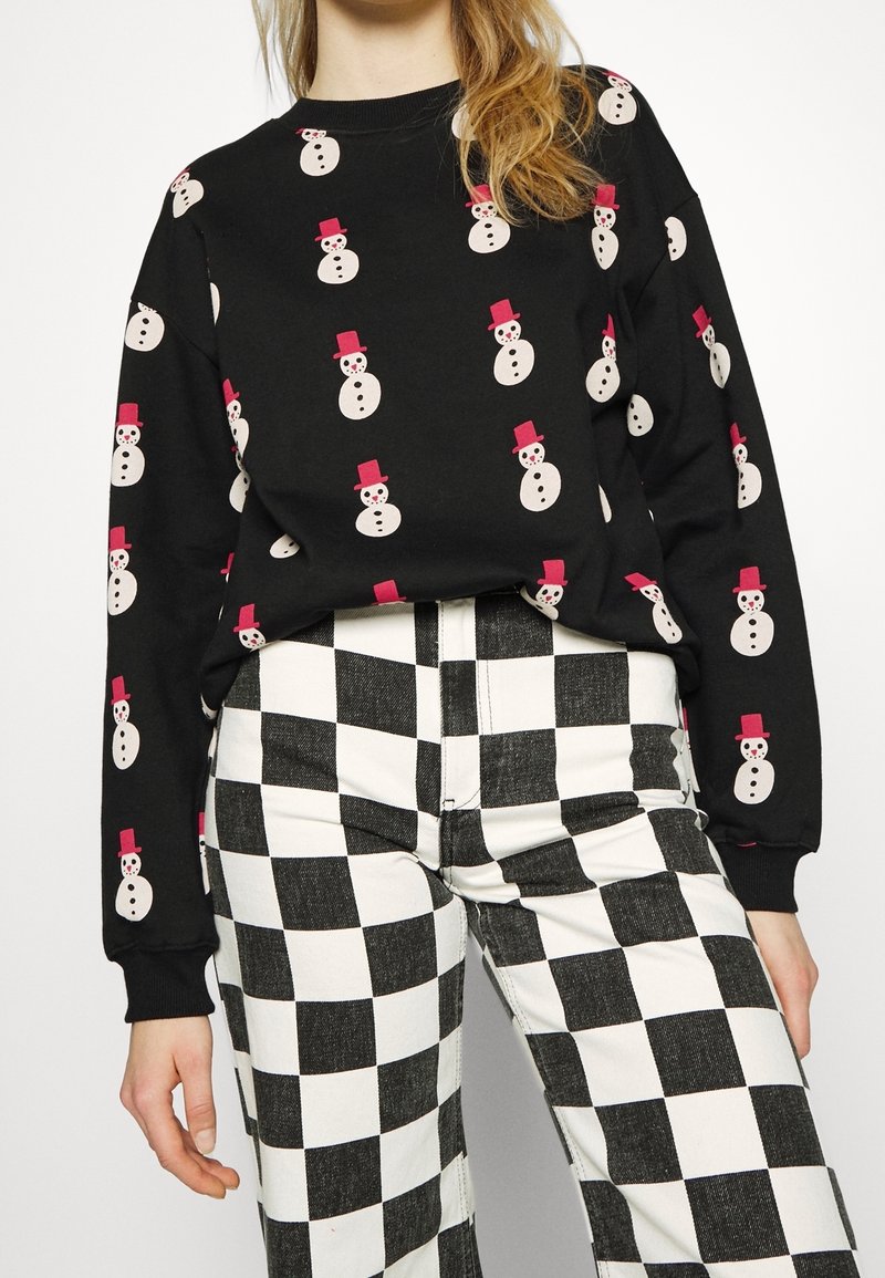 Femme portant un pull noir à motifs de bonhommes de neige et un pantalon à carreaux noir et blanc sur fond blanc uni.