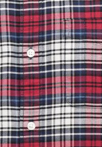 Piros, fehér és kék kockás ing, texturált anyagból készült, fehér gombokkal és keresztben futó vonalakkal, amelyek kockás mintát alkotnak.