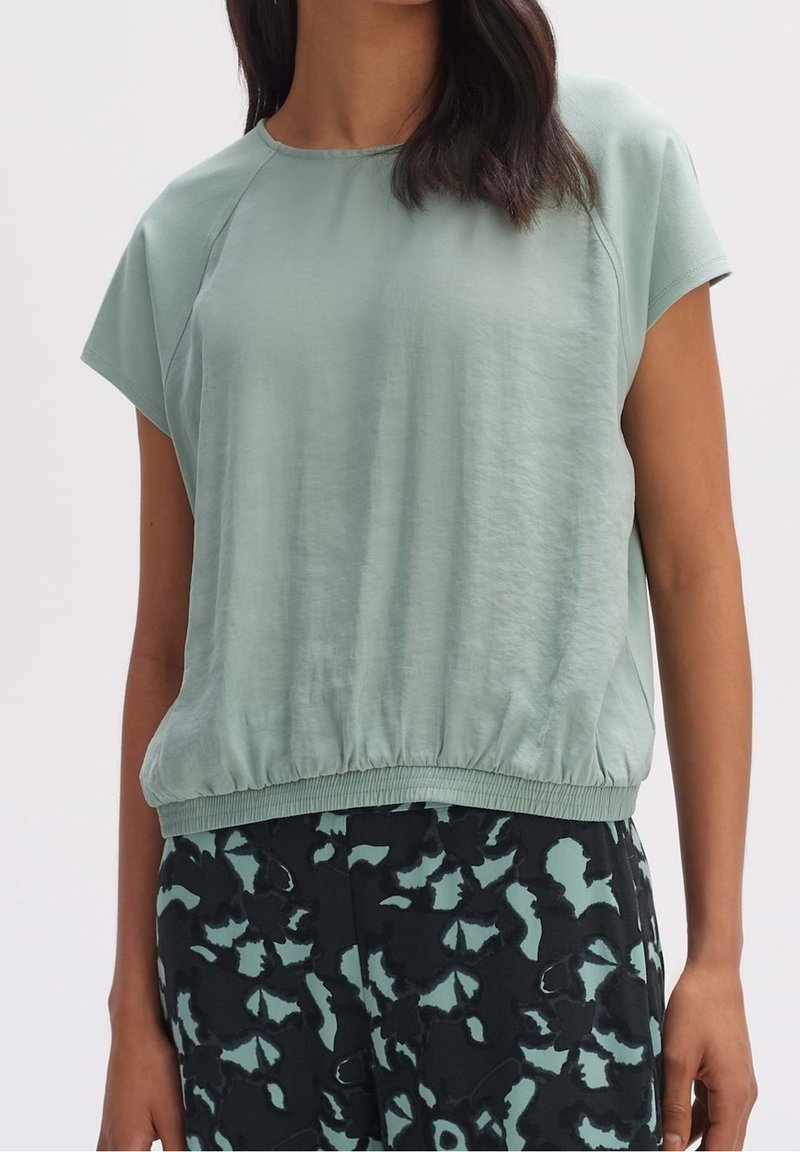 Jasnozielony top z krótkim rękawem o luźnym kroju i elastycznym dole, zestawiony z wzorzystymi ciemnymi spodniami w odcieniach zieleni i czerni.