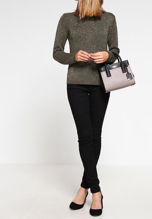 Femme portant un pull métallisé olive, un pantalon skinny noir, des talons noirs, tenant un petit sac à main bicolore au niveau de la hanche.