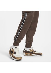 Nike Air Max sneakers med en krämfärgad ovandel, bruna accenter och en svart swoosh. Kombinerat med bruna joggers med vita sidostripes.