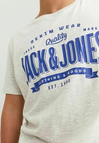 Grå kortærmet t-shirt lavet af bomuld med stor blå grafisk tekst "JACK & JONES" og yderligere branddetaljer. Blød struktur.