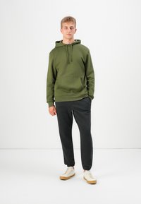 Olive green hoodie with a kangaroo pocket, paired with dark gray sweatpants. Model wears white sneakers with a light tan sole. Minimalist design.