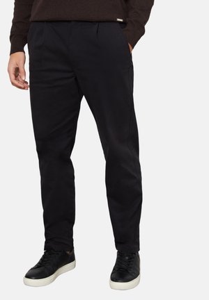 Homme portant un pantalon noir slim et des baskets noires à semelles blanches, debout avec une main dans la poche devant un fond uni.