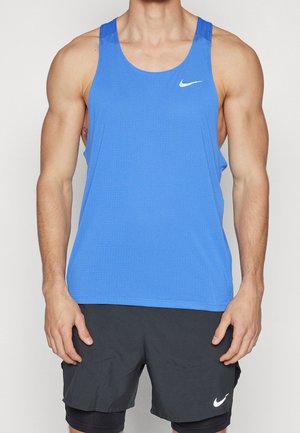 Atletyczna osoba ubrana w niebieski top Nike i czarne krótkie spodenki Nike, stojąca z rozluźnionymi ramionami wzdłuż ciała na jasnym jednolitym tle.