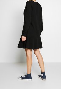 Robe noire à manches longues avec un ourlet évasé, confectionnée dans un tissu doux. Associée à des baskets montantes bleues, affichant un style décontracté.