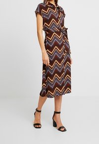 Robe à manches courtes et longueur genou, boutonnée, de couleur marron avec un motif en zigzag en or et bleu. Comprend une ceinture à nouer et un tissu texturé.
