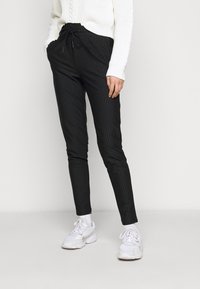 Černé kalhoty s jemným proužkem a stahovací šňůrkou v pase, úzký střih a měkká tkanina, v kombinaci s bílými teniskami. Textura vypadá hladce a stylově.