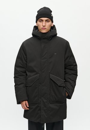 Parka nera imbottita con colletto alto, grandi tasche frontali e cappuccio. Realizzata in materiale sintetico liscio. Indossata con un berretto nero a coste.