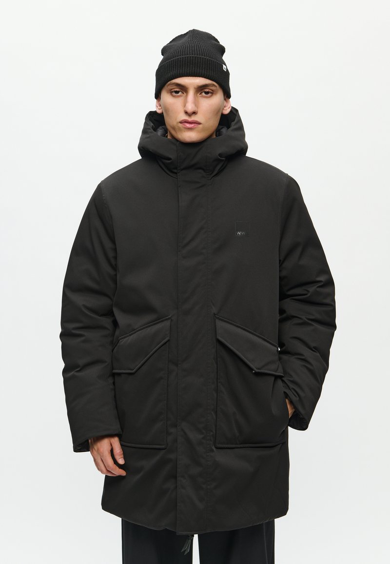 Black padded parka with a high collar, large front pockets, and a hood. Made of smooth, synthetic material. Worn with a black ribbed beanie.