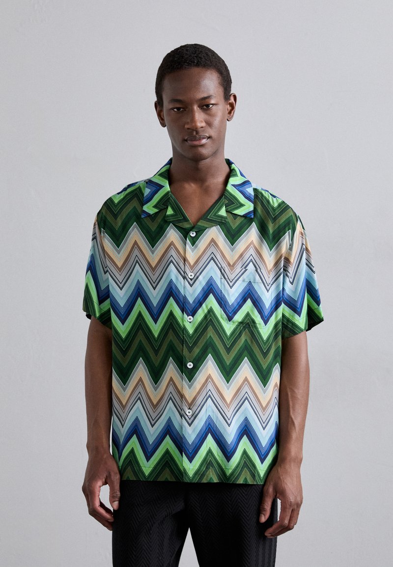 Chemise à manches courtes, à boutons, avec un motif zigzag multicolore dans des tons de vert, bleu et beige. Fabriquée en tissu léger avec une texture lisse.