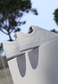 Par de zapatillas blancas de caña baja expuestas en un pedestal blanco al aire libre con árboles y cielo despejado de fondo.