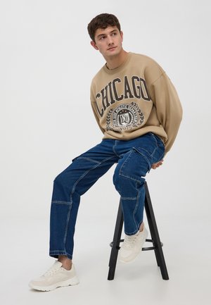 Felpa beige con grafica "CHICAGO", abbinata a pantaloni cargo in denim blu e sneakers bianche, seduta su uno sgabello nero contro uno sfondo neutro.