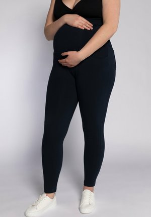 Navyblaue Umstandsleggings aus dehnbarem Stoff mit hoher Taille und zwei Seitentaschen, kombiniert mit weißen Sneakers.