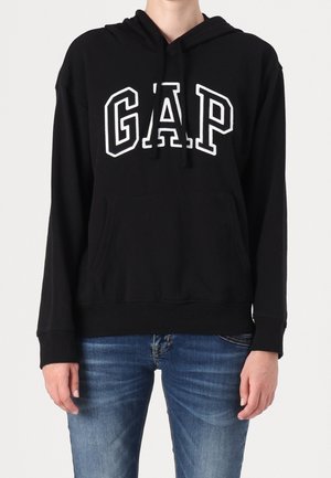 Osoba w czarnej bluzie z kapturem z białym napisem "GAP" i niebieskich jeansach, stojąca na tle jednolitego jasnego tła.