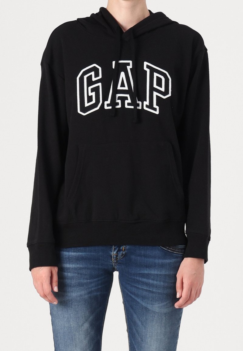 Personne portant un sweat à capuche noir avec le lettrage blanc "GAP" et un jean bleu, debout devant un fond clair uni.