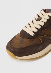 Καφέ αθλητικό παπούτσι με σουέντ επάνω μέρος, υφασμάτινη επικάλυψη με σχέδιο και λογότυπα, κρεμ κορδόνια και ελαφριά λαστιχένια σόλα με καφέ εξωτερική σόλα.