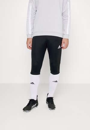 Pantalones cortos deportivos negros con el logo de Adidas en blanco, combinados con calcetas blancas hasta la rodilla que tienen acentos en negro, y botas de fútbol negras.