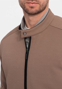 Lysbrun zip-up jakke med et struktureret mønster, høj krave og sort lynlås. Har en grå inderside og et trykknaplukning ved halsen.