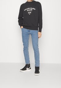Svart sweatshirt med vit "Calvin Klein Jeans 1978" logotyp, tillsammans med ljusblå slim-fit jeans och svarta högstövlar med vita accenter.