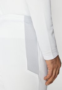 Hvit langermet topp med et grått meshpanel på siden, med teksturert stoff. Ermet avsluttes med en tettsittende mansjett.