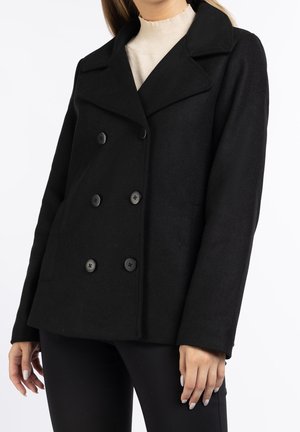 Kobieta ubrana w czarny dwurzędowy płaszcz typu pea coat na beżowym swetrze z wysokim kołnierzem i czarnych spodniach, stojąca na białym tle.