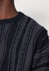 Lähekuva henkilön kaulasta ja olkapäästä, jolla on tummansininen ja harmaa neulepaita, jossa on pystysuunnassa olevia rakenteellisia kuvioita.