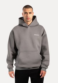 Sudadera con capucha gris, con bolsillo tipo canguro en el frente, puños y dobladillo acanalados. Presenta el texto blanco "Smilodax Sport" en el pecho. Tejido suave.