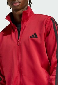 Sarkans zip-up sporta jakas izgatavots no gludas auduma, ar melnām svītrām pa piedurknēm un melnu logo kreisajā krūts apvidū.