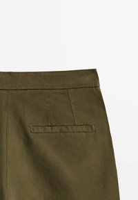 Détail d'une jupe verte olive montrant la ceinture, les coutures et une seule poche passepoilée horizontale à l'arrière sur un fond uni.