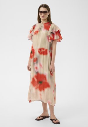 Femme portant une robe midi beige à imprimé floral rouge avec des manches courtes à volants, lunettes de soleil foncées et sandales noires, se tenant sur un fond blanc.